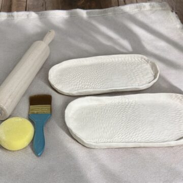 Photograph of two small white ceramic serving dishes handmade. On the left of the dishes are a brush, sponge, and rolling pin.